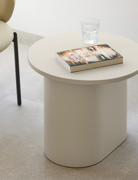 Imagen de categor&iacute;a Mesas, con mesa auxiliar redonda lacada en color beige y base robusta redondeada, junto a silla y con un libro y un vaso encima.